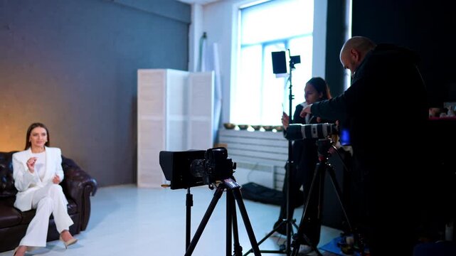 Film Crew Preparing Interview Recording In Studio. Film crew sets cameras and LED lights for seated woman interview in studio with couch and grey wall.