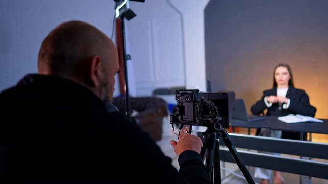 Videographer adjusting camera settings on tripod for studio interview. Over the shoulder view of camera operator setting up equipment for female presenter at desk.