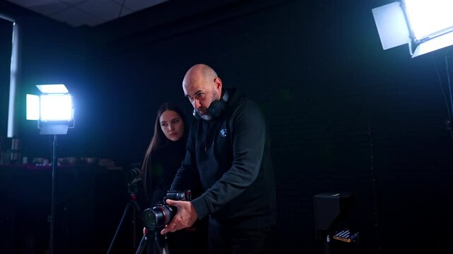 Professional videographer adjusting camera settings on tripod in studio. Middle aged bald man with headphones operating DSLR camera on tripod with assistant in dark studio.