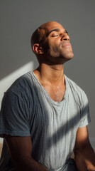 Obraz premium Serene young man with closed eyes enjoying peaceful sunlight indoors, wearing a casual grey t shirt. Calm expression and soft natural lighting create a tranquil, mindful atmosphere