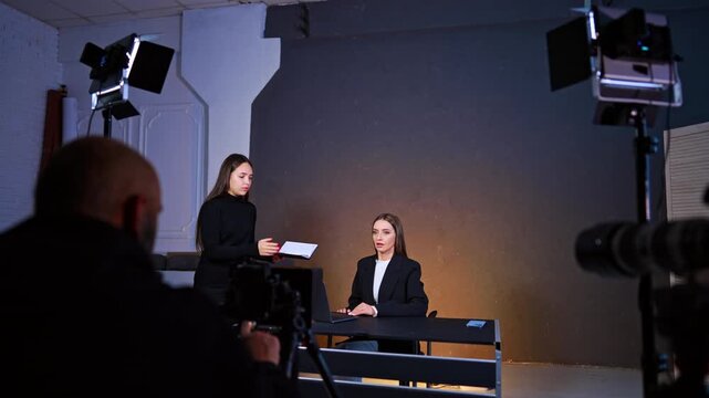 Assistant handing notebook to woman sitting at studio desk. Two women in professional attire during video shoot with lighting equipment and laptop on table in studio.
