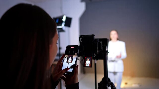 Vertical mobile phone recording of woman in white suit. Close up of smartphone screen recording video of woman standing in studio with professional equipment background.