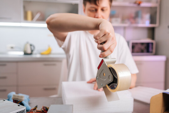 Close-up on hand sealing white box with tape dispenser. Man packs computer parts in home kitchen. E-commerce, small business, moving shipping concept.