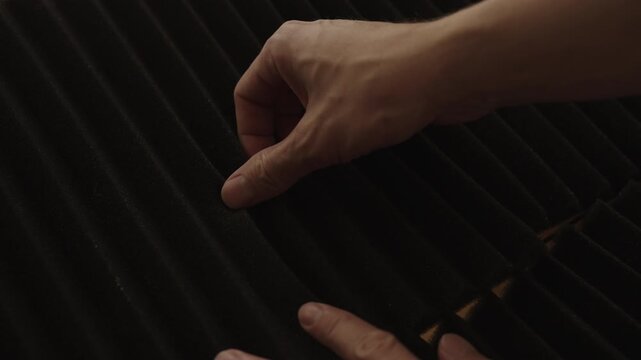 Close-up of male hands gently touching and stroking textured surface of black acoustic foam panel, exploring material used for soundproofing and sound absorption in recording studio, slow motion.