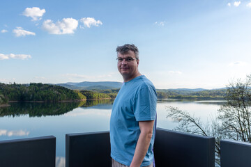 A middle aged man in a blue shirt stands at a scenic lake viewpoint with calm water, forested hills, mountain ridges, blue sky, and reflections in the background, creating a travel and outdoor lifesty © Volha