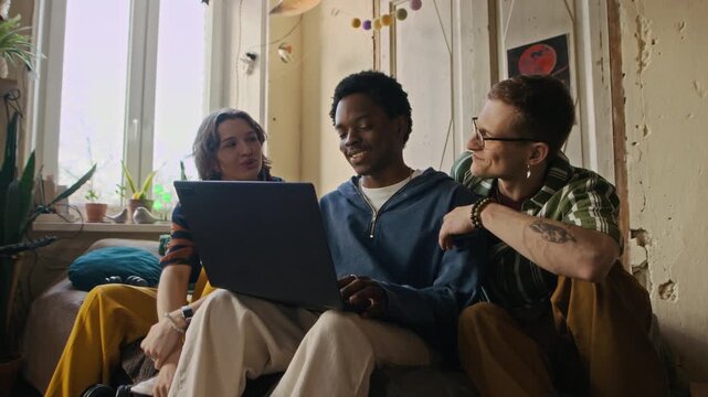 Medium long shot of young African American freelancer refusing to hang out with two Caucasian roommates and continuing working on laptop in shared apartment
