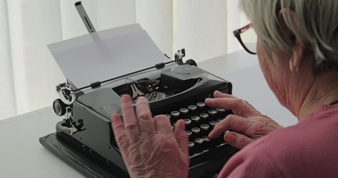 Over-the-shoulder view of a senior woman pressing the keys on a vintage black manual typewriter with paper loaded