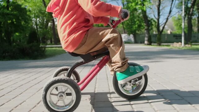 A child rides a three-wheeled bicycle through a park in summer, the camera zooming in on the spinning wheels and pedals from a low angle.