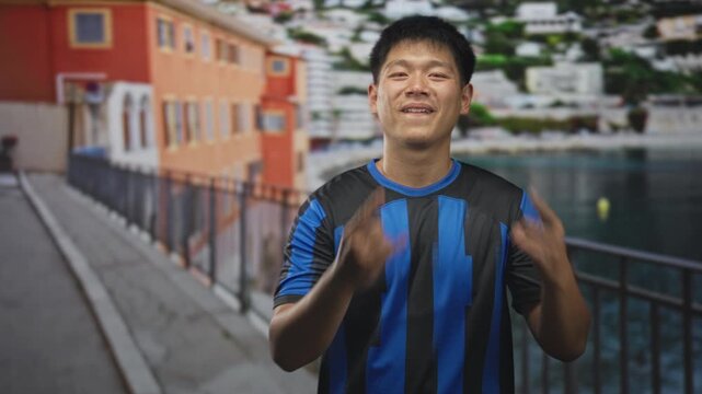 Man in blue and black soccer jersey makes rock horns with bare hands on a street by the waterfront; playful confidence.