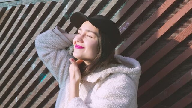 Beautiful girl in a baseball cap and fluffy jacket closing eyes from bright sunbeams.