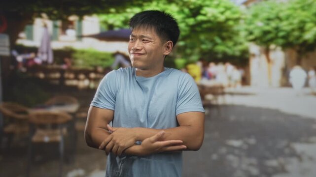 Young chinese man arms crossed hugging chest in street cafe with outdoor tables, umbrellas and leafy trees; unease vulnerability.