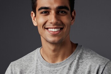 Obraz premium Handsome man smiles warmly showing white teeth. He looks directly at camera with confident, happy expression. Gray shirt contrasts with tanned skin in studio portrait.