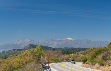 Fototapeta premium A car travels along the winding Rim of the World Scenic Byway (State Route 18), with a panoramic view of the snow-capped Mount San Antonio (Mount Baldy) in the distant San Gabriel Mountains