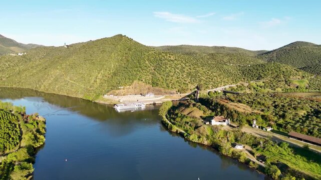 Aerial view of Barca de Alva on the Douro River with a cruise boat docked near lush terraced hills and clear blue sky in Figueira de Castelo Rodrigo, Distrito da Guarda, Portugal.