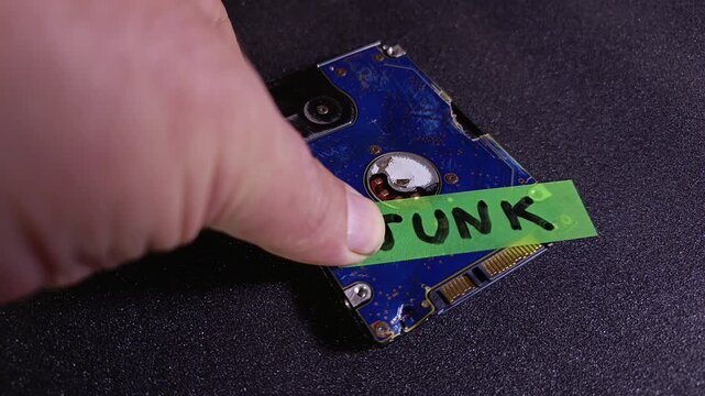 Person's hand sticking a junk green note onto damaged hard drive with dark background. Concept of AI model collapse, corrupted data, e-waste recycling, system failure, broken and outdated equipment.
