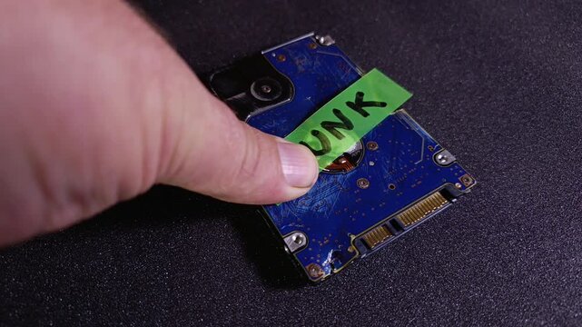 Person's hand sticking a green note to damaged hard drive with junk written on note. Concept of AI model collapse, corrupted data, e-waste recycling, system failure, broken and outdated equipment.