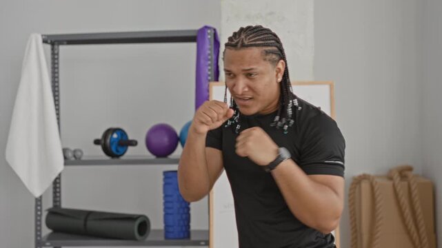 Man throwing a jab with fists raised, braided hair, wearing black shirt, visible forearm and smartwatch near foam roller and battle rope in a studio; training determination.