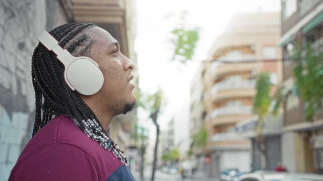 Man touching headphones with hand to ear on street near apartment building and graffiti wall; urban listening serenity.