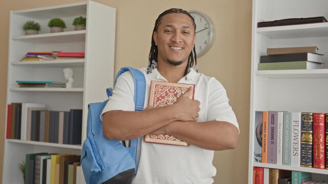 Man holding ornate hardcover book against chest with both arms, backpack on shoulder, smiling in building library with bookshelves and clock visible; academic confidence.