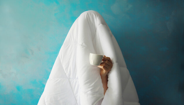 Lazy woman waking up after sleeping covered with white blanket holds cup of coffee in the morning