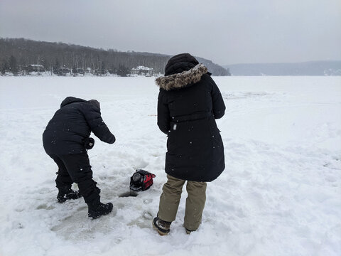 A man and woman in heavy winter gear stand over an ice hole, using a portable fish finder on a vast snow covered lake with a forested shoreline in the distance.
