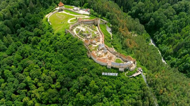 Aerial view of Rasnov Citadel surrounded by dense green forests and the Rasnov sign on the hillside in Rasnov, Brasov, Romania.