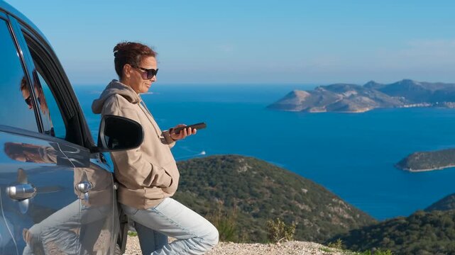 Woman using a smartphone on a scenic road trip overlooking the sea. Casual woman leaning against her car and using a smartphone while enjoying the beautiful sea view with islands during road trip