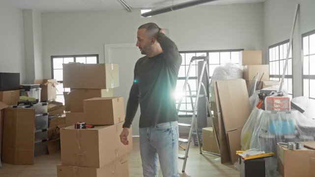 Man with hand on neck beside stacked cardboard boxes and a ladder in a building during unpacking and setup of a new home; moving fatigue.