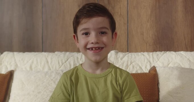 Portrait of laughing 8 year old boy sitting on sofa in living room at home, looking at camera. Natural childhood stage with missing baby teeth, cozy home atmosphere, lifestyle concept.