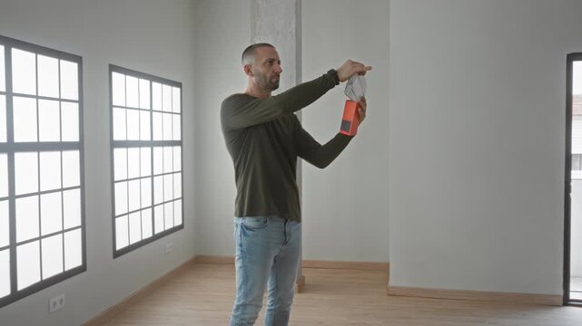 Young hispanic man fans paint swatches and holds a red sample with bare hand in building; color testing contemplation.