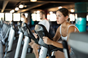 Fototapeta premium Girl spends time in gym, improves physical shape with exercises. Female sport studio hall visitor uses exercise bike to pump leg muscles. Strength and motivation, sport, fitness goal