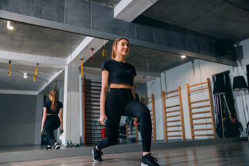Obraz premium Young woman exercising with dumbbells during gym lunge workout. Attractive woman training legs inside modern indoor fitness studio. Strength sport concept.