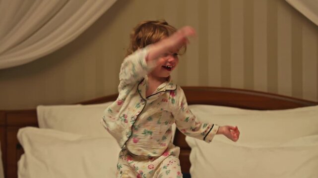 Joyful little girl wearing pajamas with curly hair happily jumping and dancing on a large bed. Carefree childhood concept showing a toddler having fun in the bedroom before going to sleep