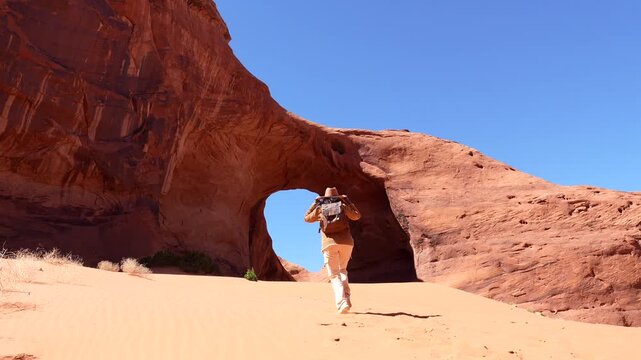 Happy tourist woman with a backpack walking on sand dunes toward a natural red rock arch, enthusiastically throwing her hat in the air at monument valley tribal park in arizona