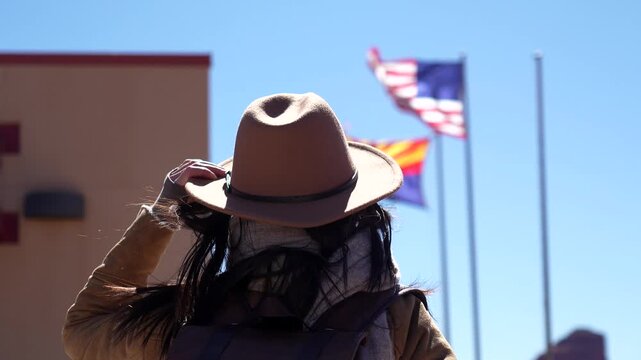 Young adventurous woman wearing a brown cowboy hat looking at the american and arizona flags waving in the wind in monument valley, a famous landmark on the border of arizona and utah
