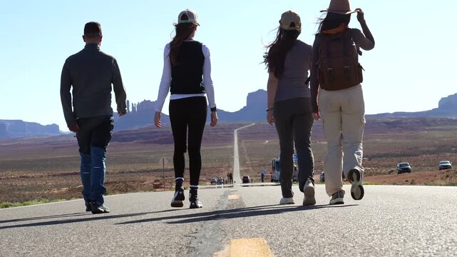 Young friends walking away on the famous desert road leading to monument valley, with one woman waving her hat in a gesture of freedom and happiness during a scenic american road trip