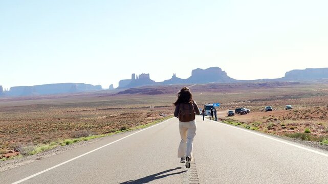 Energetic female traveler with a backpack happily running down the iconic highway at forrest gump point, enjoying her freedom on a scenic road trip through monument valley national park