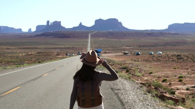 Young female traveler with a backpack and hat walking on a scenic desert highway towards monument valley at forrest gump point, embarking on an adventurous journey through the american west