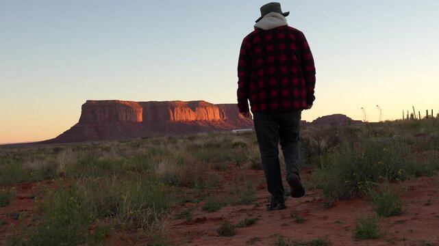 Hiker with a plaid shirt walking away from the camera through the desert during a beautiful sunrise, with the iconic sandstone buttes of monument valley glowing in the background