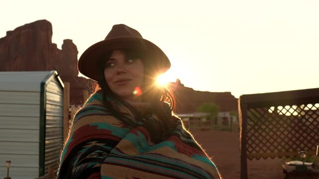 Beautiful young woman wrapped in a colorful native american pattern blanket, wearing a hat and enjoying the sunrise with the stunning rock formations of monument valley in the background