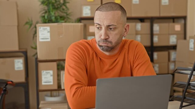 Man checking watch at laptop amid stacked cardboard boxes in parcel office building wearing orange sweater and looking at screen; frustration deadline pressure.