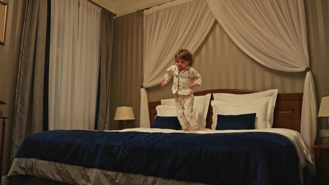 Energetic toddler wearing pajamas joyfully jumping and bouncing on a large canopy bed in a cozy hotel room, expressing happiness and carefree childhood fun before going to sleep