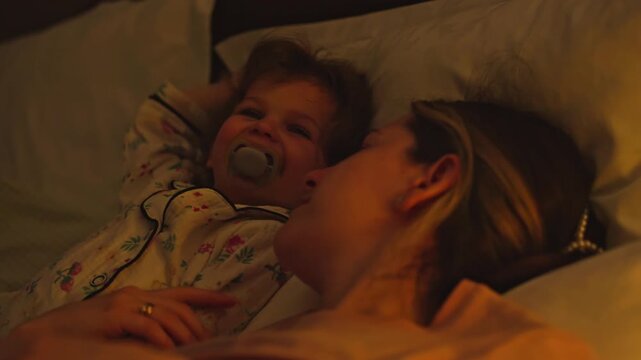 Affectionate mother comforting her crying toddler daughter in bed before sleep, giving a gentle kiss to soothe her. Loving family moment showing maternal care, tenderness, and bedtime routine