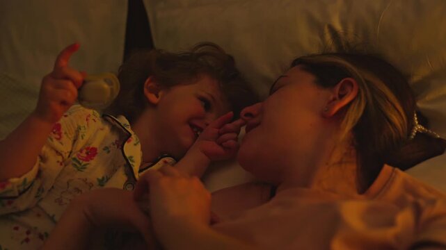 Little girl in pajamas playing and laughing with her loving mother while lying together in bed at night. Sweet family moment of tenderness and affection before falling asleep
