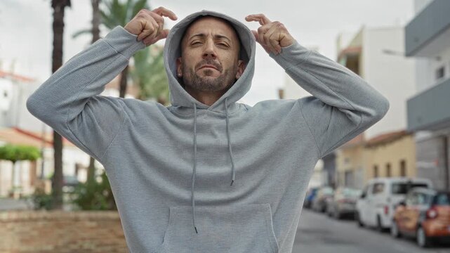 Man with face visible pulls and adjusts hoodie while crossing arms and looking sideways on a palm lined street with parked cars; thoughtful resilience.