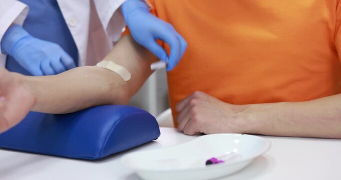 Doctor in medical gloves applying adhesive bandage onto patient's arm after drawing blood sample at white table in clinic, closeup. Camera moving right