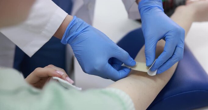 Doctor in medical gloves applying adhesive bandage onto patient's arm after drawing blood sample at white table, closeup. Camera moving down