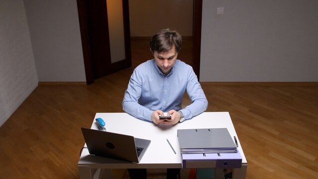Young man in a home office engrossed in his smartphone, scrolling social apps and ignoring laptop and documents while procrastinating work and losing focus on tasks