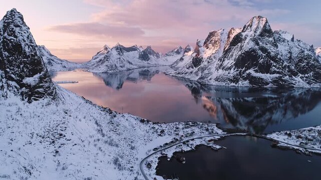 Aerial view of the Lofoten islands with snow-capped peaks reflecting in the calm fjord waters during a pink sunset in Reine, Nordland, Norway.