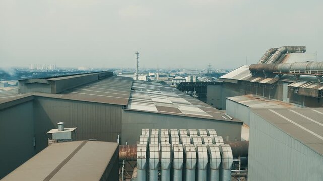 Cinematic aerial descending shot of a large industrial factory complex featuring ventilation systems and city skyline background.